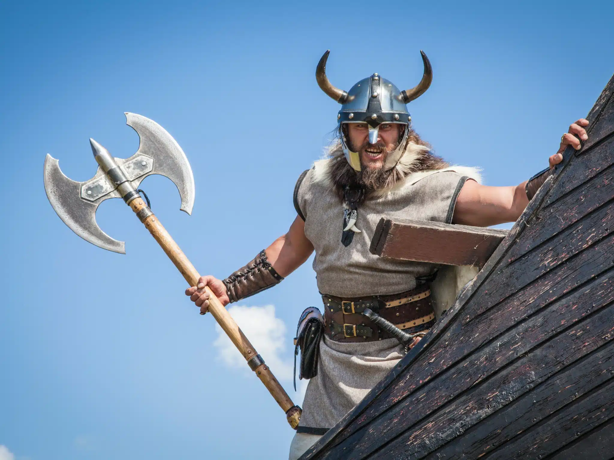 Viking avec casque à cornes sur pont de drakkar, hache à la main, sous ciel bleu.