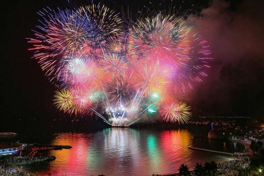 Feux d'artifice éclatent sur l'eau, foule émerveillée au bord, ambiance festive nocturne.