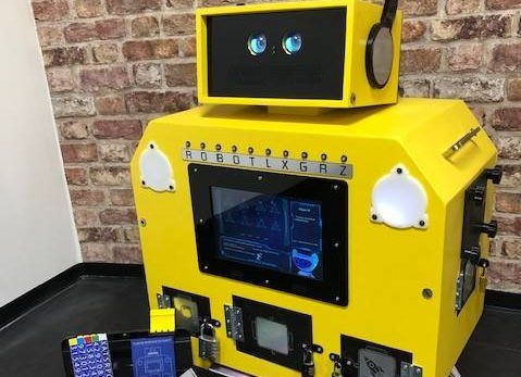 Grand robot jaune interactif, boutons numérotés, devant un mur de briques avec jeux d'escape colorés.