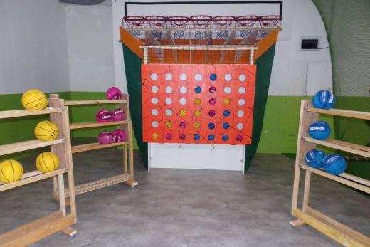 Terrain de basket ludique, panier vertical, ballons colorés rangés sur les côtés.