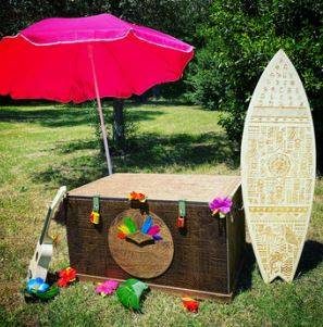 Coffre fleuri, ukulélé, planche et parasol rose sur l'herbe. Ambiance plage festive et colorée.