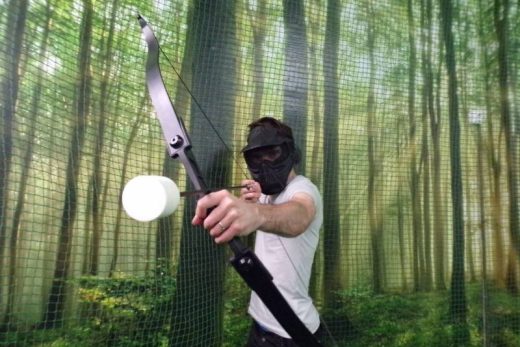 Déguisé, un joueur vise à l'arc dans une salle décor forêt, filet en fond, ambiance immersive.