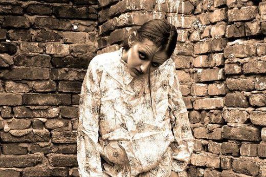 Personne en chemise à motifs devant un mur de briques, tête inclinée, regard baissé.