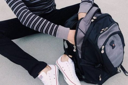 Personne assise au sol, chemise rayée, pantalon noir, baskets blanches, ouvre un sac à dos noir et gris.