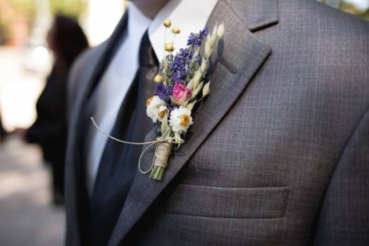 Costume gris, cravate noire, boutonnière de fleurs colorées épinglée au revers.