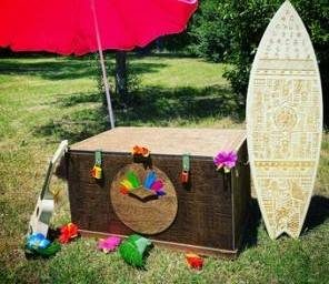 Coffre fleuri, surf décoré et ukulélé sous grand parasol rouge en plein air, ambiance estivale.