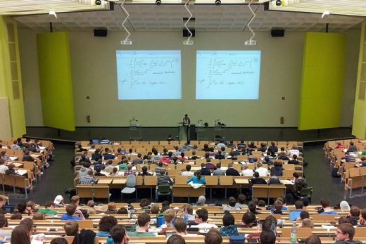 Amphithéâtre bondé, étudiants écoutant un professeur devant deux écrans présentant des équations mathématiques.