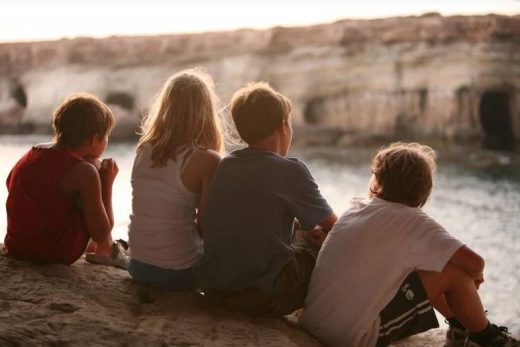 Quatre enfants sur un rocher face à la mer, falaises et grottes visibles en arrière-plan.