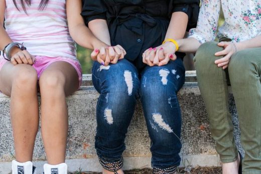 Sur un banc, trois personnes assises ; deux au centre se tiennent la main. On voit jambes et torses.