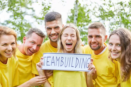Équipe de six en jaune pose dehors, pancarte TEAMBUILDING, arbres et verdure en fond.
