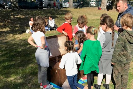 Enfants réunis autour d'un coffre lors d'un jeu immersif en plein air, soleil et spectateurs assis.