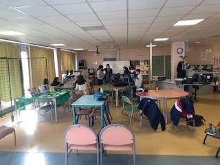 Salle lumineuse, étudiants aux ordinateurs, manteaux sur chaises, rideaux jaunes aux fenêtres.
