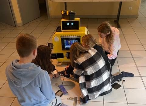 Cinq enfants jouent autour d'un robot jaune, cartes et jeux étalés sur le sol, ambiance ludique.