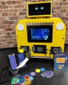 Borne robot jaune, écran d'icônes, matériel éducatif varié devant, mur en briques en arrière-plan.