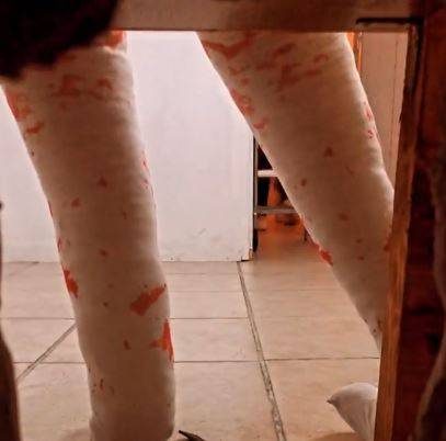 Deux jambes plâtrées tachées d'orange sous une table en bois, sol carrelé.