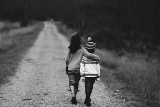 Deux enfants marchent, bras dessus bras dessous, sur un sentier entre herbe et arbres.