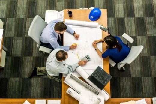 Trois personnes étudient des plans à une table, casque bleu, ordinateur portable et calculatrice posés.