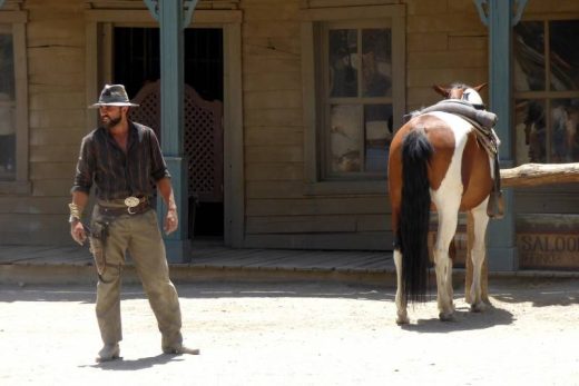 Cowboy devant un bâtiment en bois ancien, cheval sellé à proximité, ambiance western immersive.